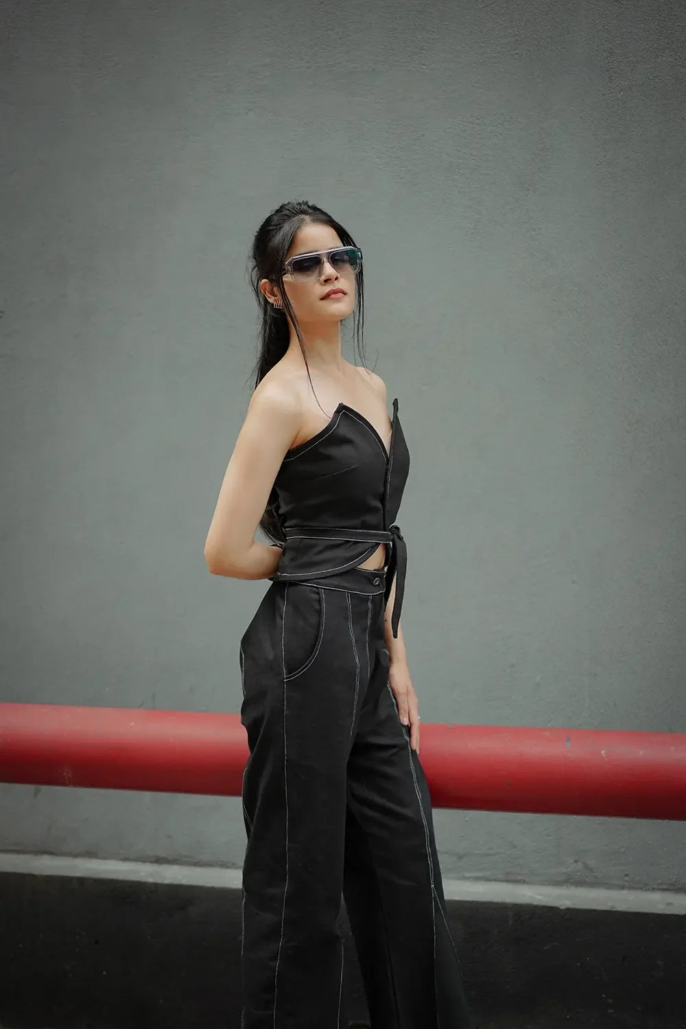 Woman in black sleeveless top and pants, wearing sunglasses, standing by a gray wall, Streiq fashion