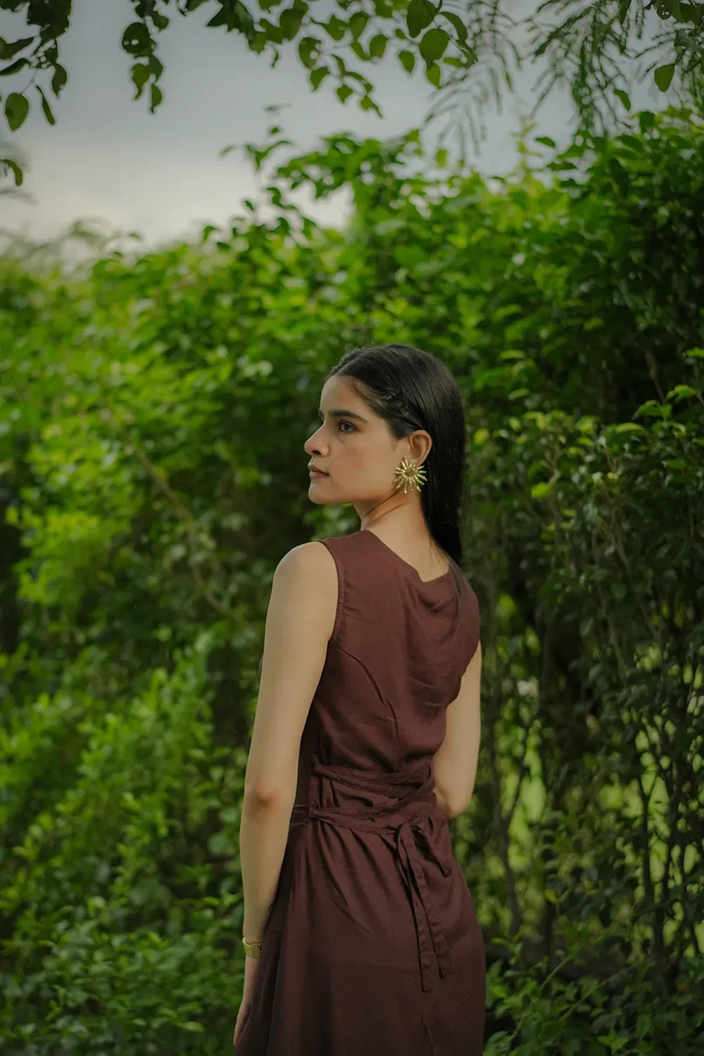 Woman in a brown sleeveless dress with gold earrings standing outdoors near lush greenery, Streiq