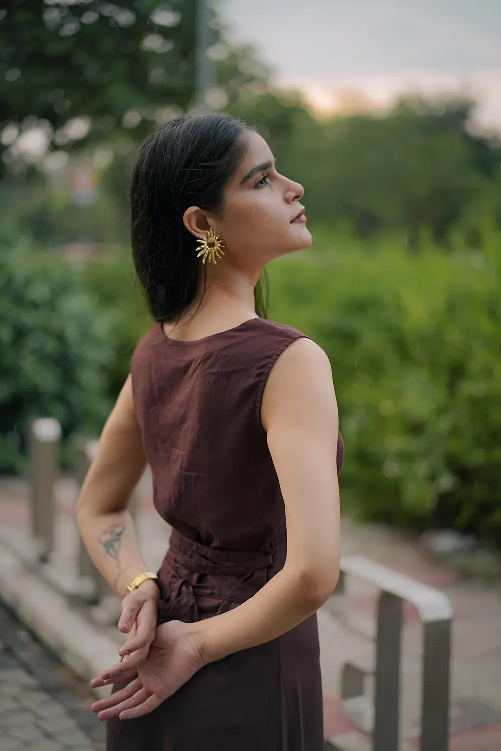Woman in a sleeveless brown dress with gold sunburst earrings outdoors, Streiq fashion