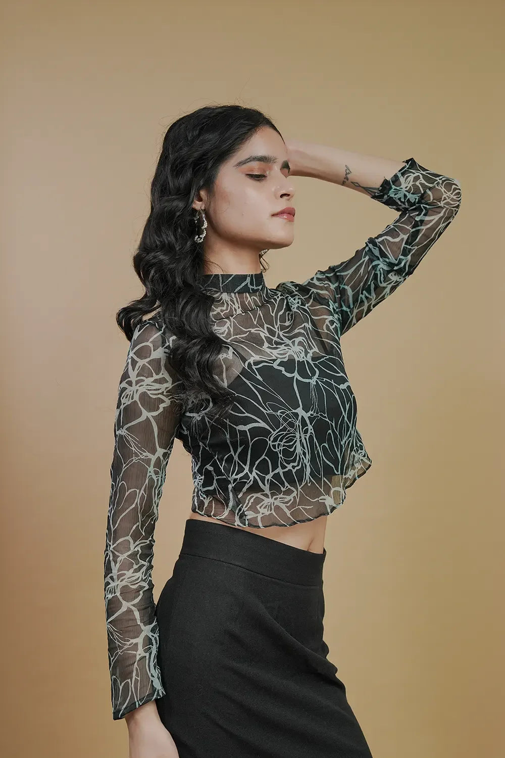 Woman in sheer floral mesh long sleeve top and black skirt, posing against beige background