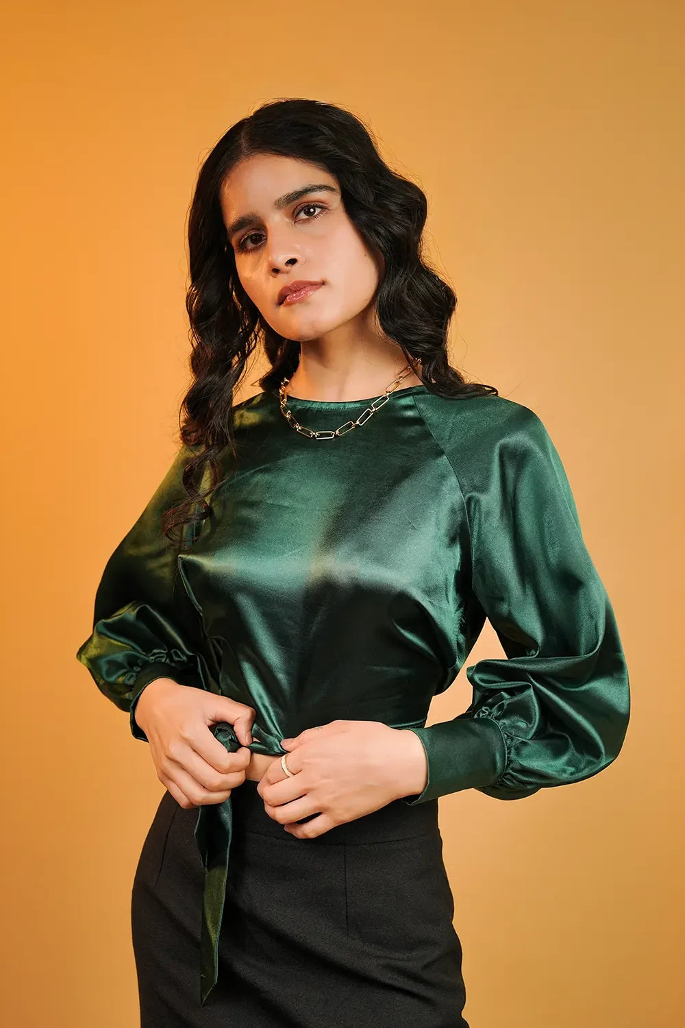 Woman in emerald green satin blouse and black skirt, posing against a warm yellow background