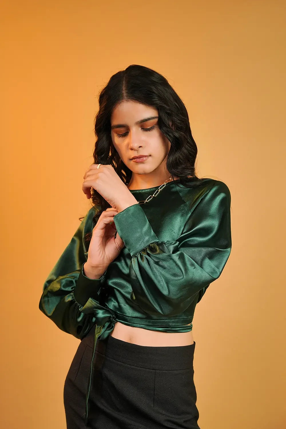 Woman in a green satin blouse and black pants posing against a solid orange background
