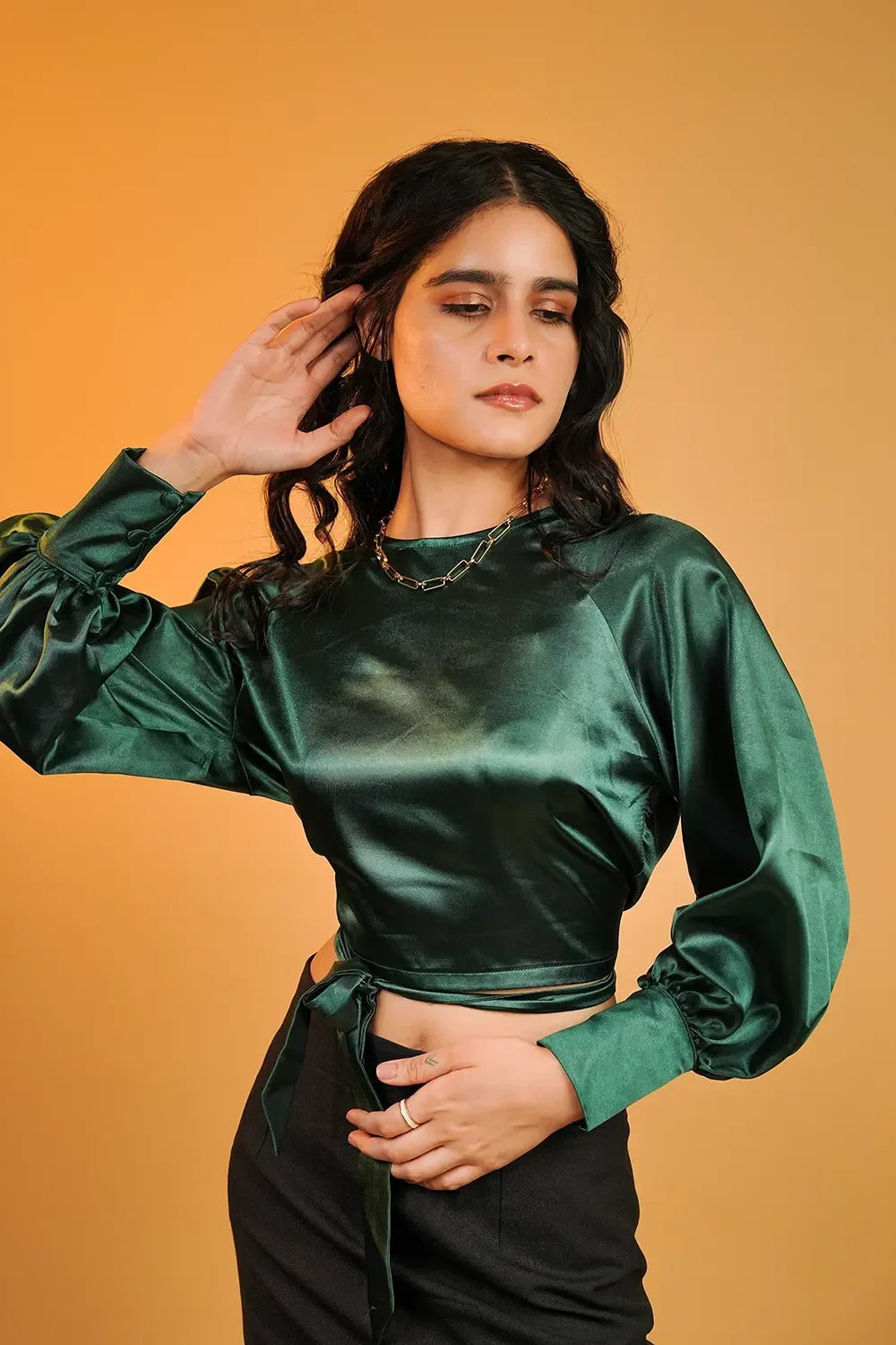 Woman in emerald green satin crop top with puff sleeves, posing against a warm yellow backdrop