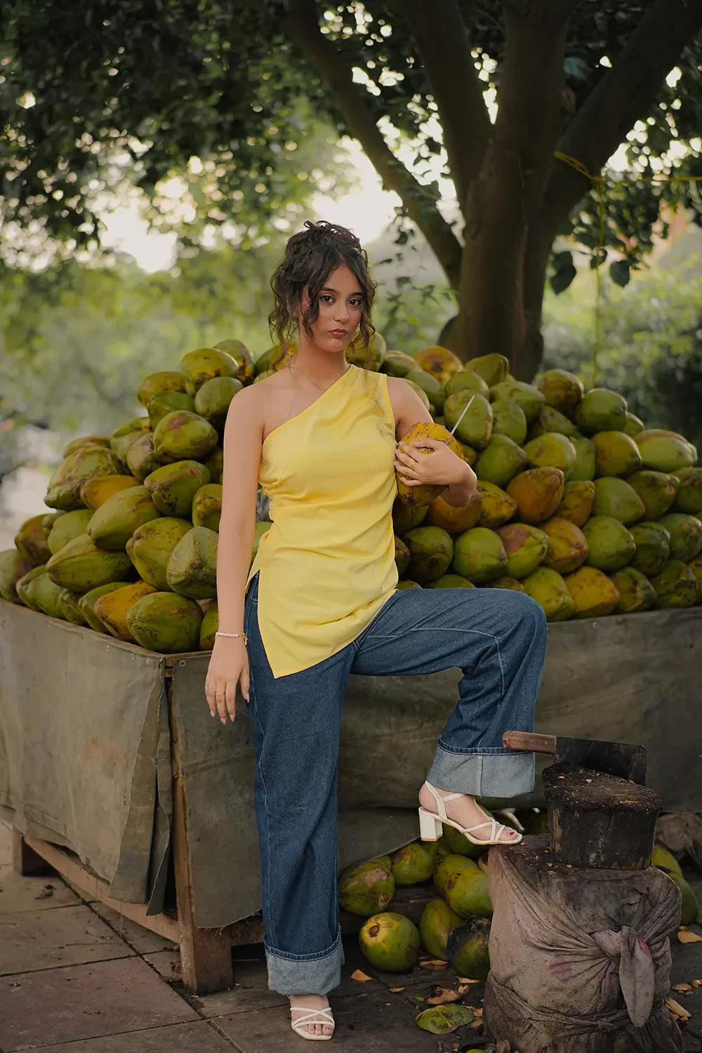 Woman in yellow one-shoulder top and jeans holding coconut at outdoor market, Streiq fashion