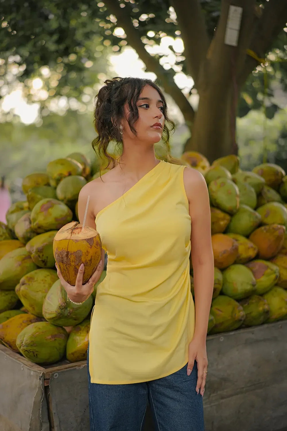 Woman in yellow one-shoulder top holding coconut, standing by fresh coconuts, Streiq style