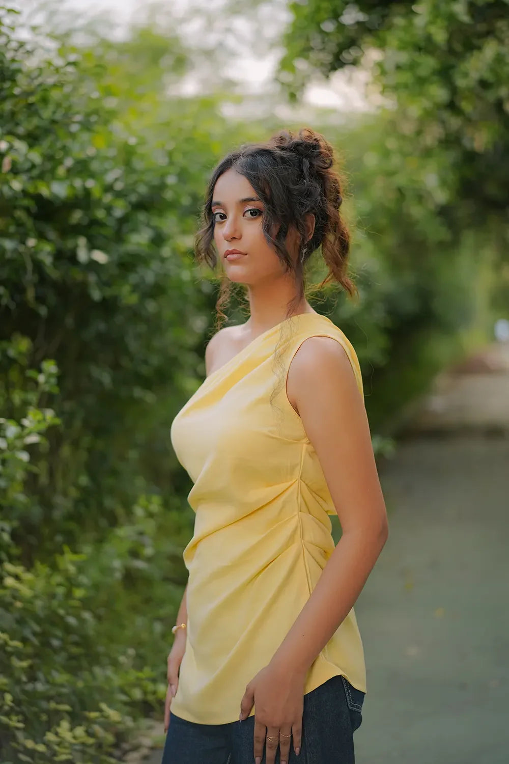 Woman in yellow sleeveless top posing outdoors, Streiq fashion, green leafy background