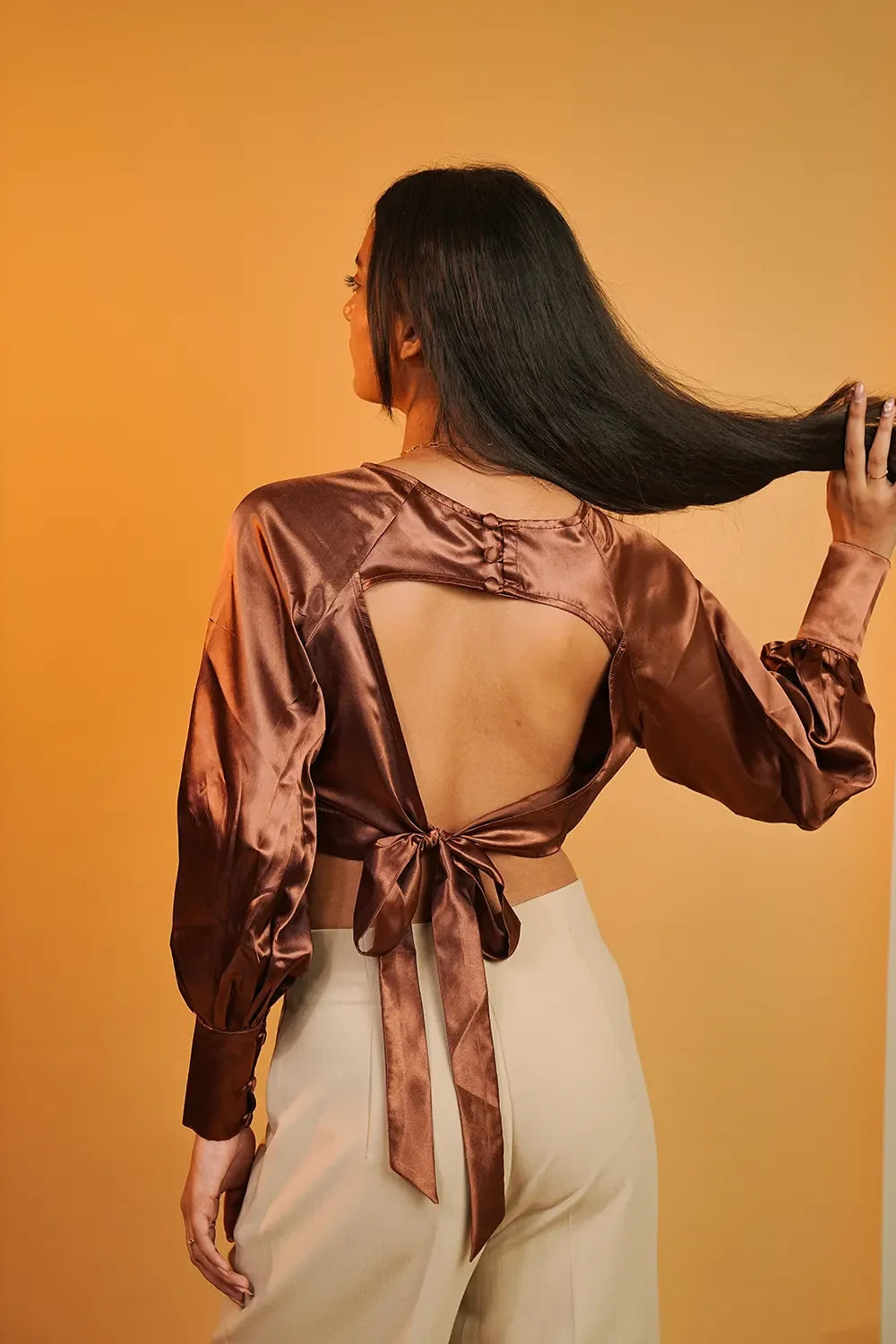 Woman in satin brown open-back top and beige pants on orange background, Streiq fashion