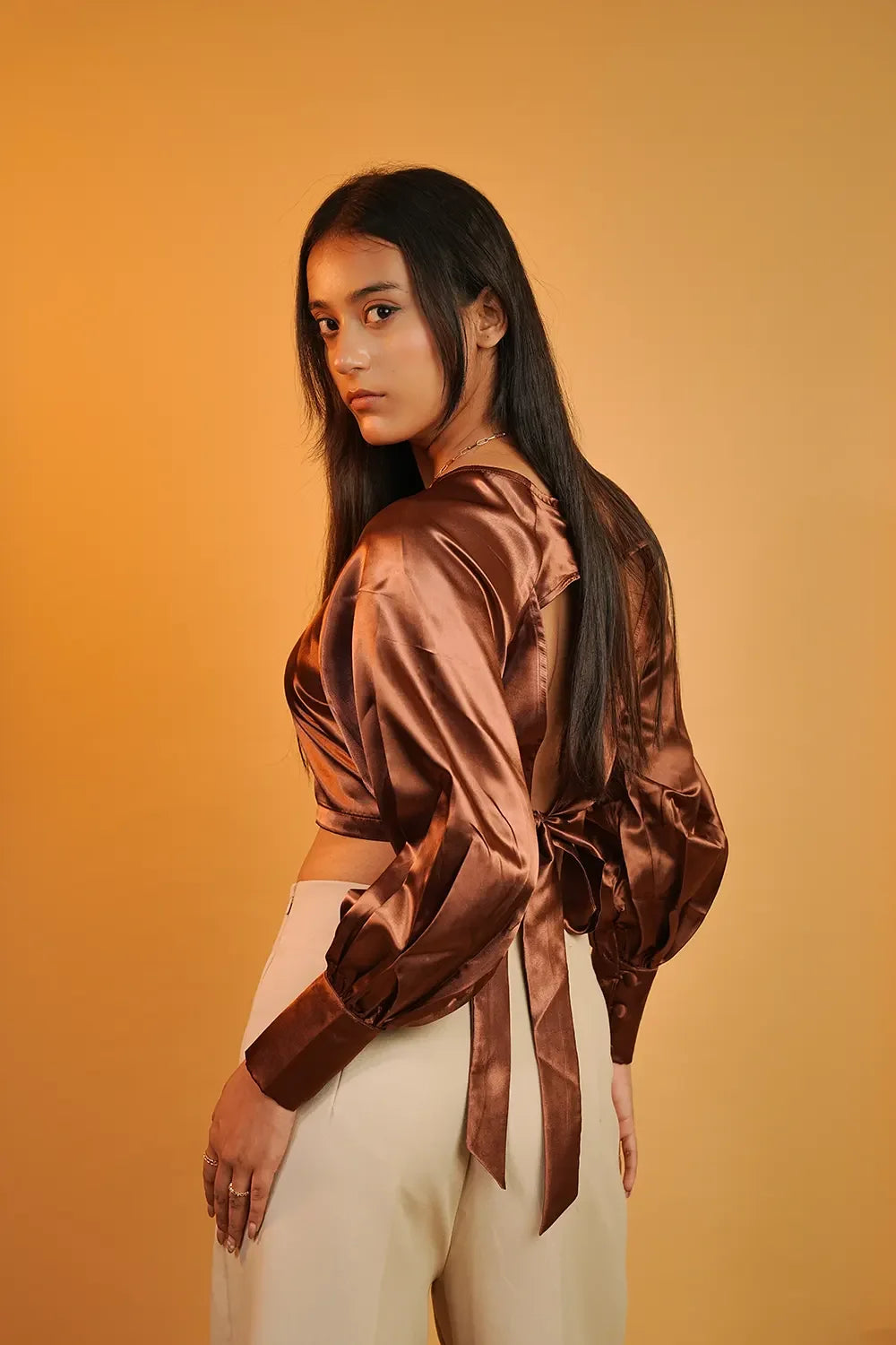 Woman in brown satin open-back blouse and cream pants against warm yellow background, Streiq fashion