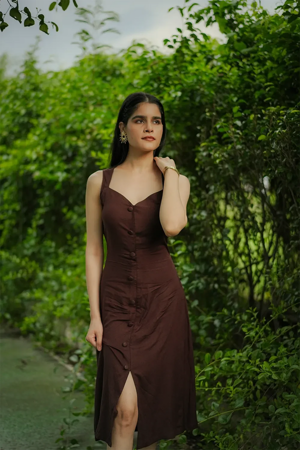 Woman in a sleeveless brown dress with button front, posing outdoors amid lush green foliage